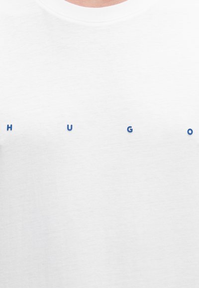 Бяла риза с малки, разположени на разстояние сини букви, изписващи "HUGO" върху гърдите.