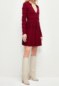 Robe tricotée bordeaux avec un décolleté en V profond et des rayures texturées, associée à des bottes hautes beiges à talons. Texture lisse et coupe ajustée.