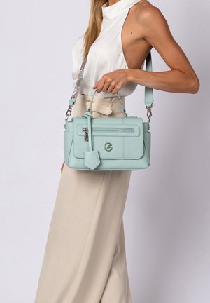 Woman wearing sleeveless white top and beige pants holding a light blue leather handbag with front zipper and detachable strap.