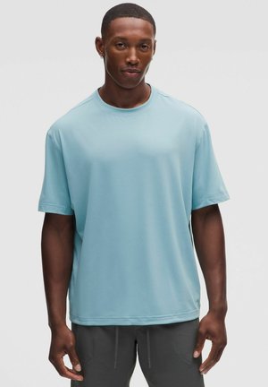 Giovane uomo con capelli corti e orecchino, che indossa una camicia azzurro chiaro a maniche corte e pantaloni scuri su uno sfondo semplice.