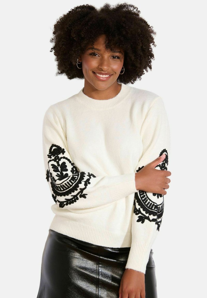 Maglione bianco con motivi ricamati neri sulle maniche, collo e polsini a coste, abbinato a una gonna di pelle nera.