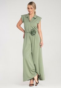 Zielony kombinezon z kołnierzykiem, bez rękawów, z guzikami z przodu oraz paskiem z kwiecistym akcentem w talii.