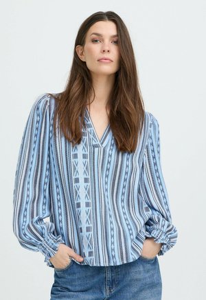 Kvinde med langt brunt hår iført en blå og hvid mønstret bluse med lange ærmer og blå jeans med hænderne i lommerne mod en ensfarvet baggrund.