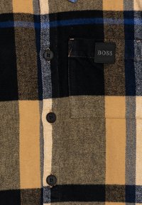 Koszula flanelowa w kratę w brązowym, czarnym i niebieskim kolorze z kieszenią na piersi, czterema czarnymi guzikami oraz naszywką z logo BOSS. Miękka tekstura i ciepły materiał.