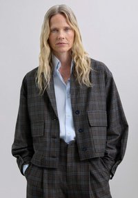 Chaqueta de gran tamaño a cuadros marrones con dos bolsillos frontales, usada sobre una camisa de botones azul claro; presenta botones azules prominentes y un corte relajado.