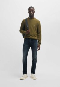 Olive green textured sweater with a round neckline, paired with dark blue jeans and beige sneakers. Backpack over one shoulder.