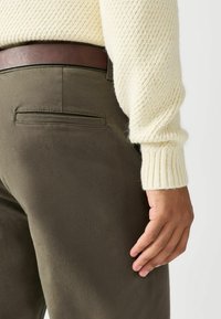 Tumedate rohelised püksid sileda tekstuuriga, tagaküljel on tasku ja pruun nahkvööt. Kanda kreemivärvi kootud kampsuniga.