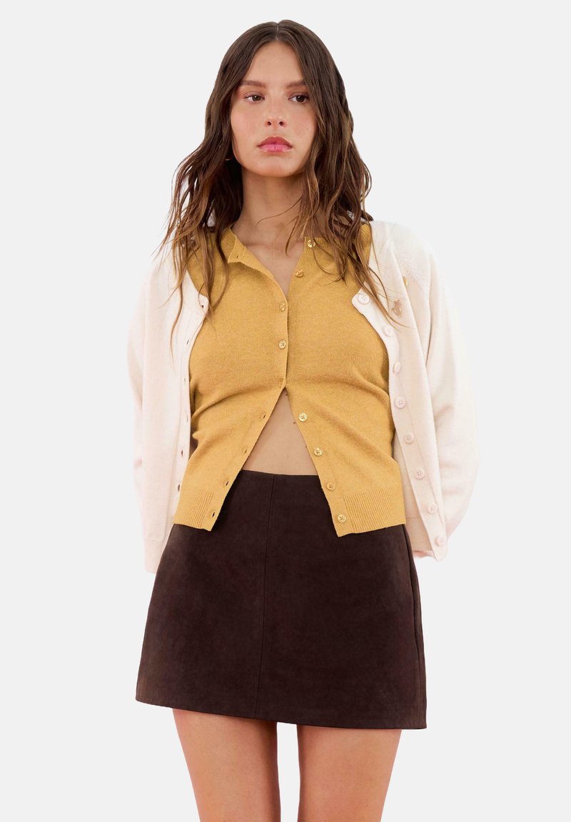 Donna con capelli castani mossi indossa un cardigan giallo senape con bottoni sopra un maglione bianco e una gonna A-line marrone scuro, in piedi su uno sfondo neutro.