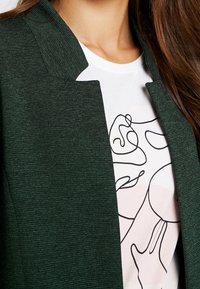 Blazer vert texturé avec un col montant, porté sur un T-shirt blanc arborant un dessin au trait noir et des formes abstraites aux tons doux.