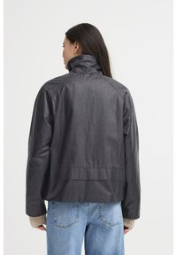Svart kort jacka med hög krage, tillverkad av mjukt tyg, med raglanärmar och en enkel bak sömdetalj. Bärs över ljusa jeans.