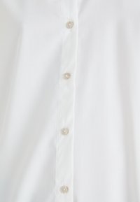 Gros plan d’un tissu blanc avec quatre boutons beiges attachés sur une patte verticale.