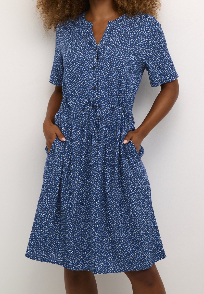 Femme portant une robe bleue mi-longue à manches courtes, avec un petit imprimé floral, une patte de boutonnage devant et des poches, debout les mains dans les poches.