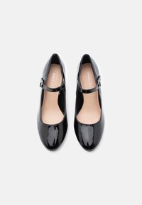Černé lakované boty Mary Jane s přezkami, kulatými špičkami a béžovými stélkami na bílém pozadí.