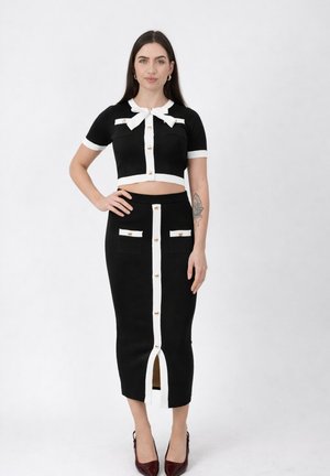 Frau trägt ein schwarzes, bauchfreies Oberteil und einen geknöpften Midirock mit weißem Rand, steht mit der Hand in der Hüfte vor einem einfarbigen Hintergrund.