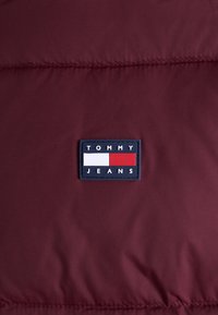 Giubbotto imbottito bordeaux con una texture liscia, dotato di una toppa rettangolare blu navy con "TOMMY" in bianco e "JEANS" in bianco su uno sfondo rosso.
