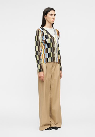 Γυναίκα που φοράει πολύχρωμο καρό ζακέτα με κουμπιά, λευκό πουκάμισο, φαρδιά μπεζ παντελόνια και μαύρα παπούτσια, στέκεται μπροστά σε απλό φόντο.