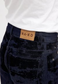 Tummansininen samettihousut, joissa on nahkainen etiketti, jossa lukee "NA-KD". Suunnittelussa on tavalliset taskut ja hillityt tekstuurivaihtelut.