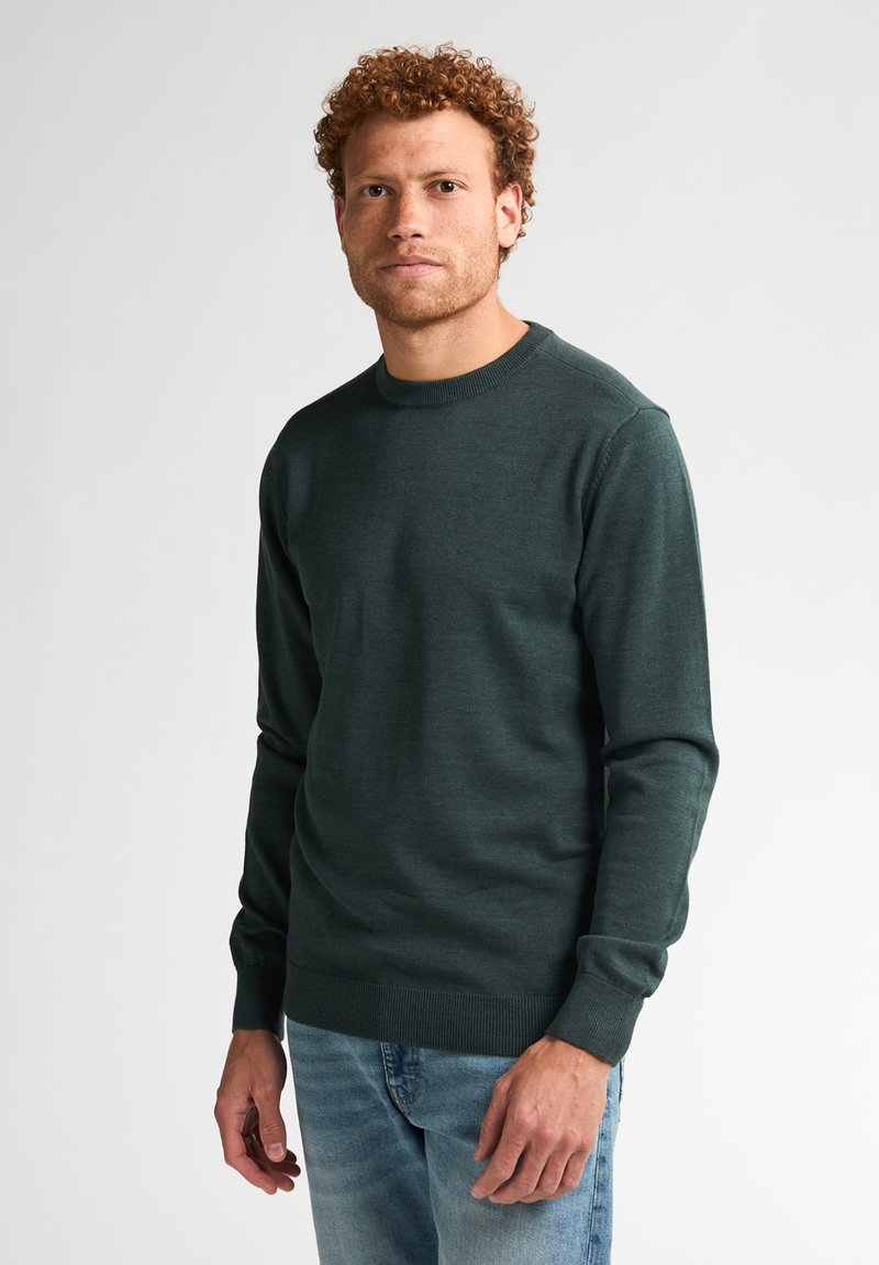 Donkergroene crewneck trui gemaakt van zachte gebreide stof. Heeft lange mouwen en ribgebreide boorden en zoom, gecombineerd met lichtblauwe jeans.