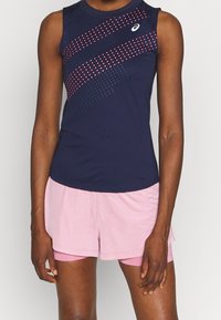 Débardeur de sport sans manches en marine avec un motif à pois en rose et bleu ; associé à un short rose clair. Le tissu semble léger et respirant.
