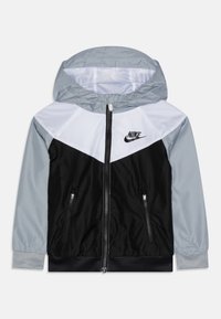 Nike bunda s šedou kapucí, černým tělem a diagonálními bílými a šedými panely. Bunda má zipové kapsy a elastické lemování. Lehký materiál.