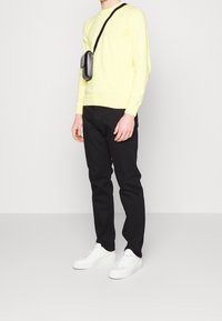 Hellgelber Pullover, schwarze Hose und weiße Sneaker. Eine kleine schwarze Umhängetasche wird getragen. Glatte Textur, einfaches Design.
