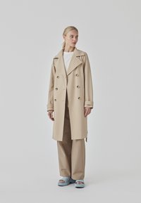 Beige double-breasted trench coat with wide lapels, large gold buttons, and rolled cuffs, paired with cream top, beige trousers, and blue platform sandals.