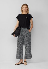 Czarny t-shirt z krótkim rękawem z detalem w kształcie gwiazdy, zestawiony z szerokimi spodniami w czarno-białe wzory. Model trzyma fakturowaną czarną torebkę.