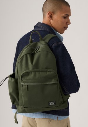 Groene rugzak met een gestructureerde afwerking, verstelbare schouderbanden, een voorvak met rits en een zijnetwerkvak. Draagt over een denim jas.