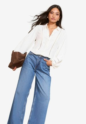 Wit overhemd met knopen, met horizontale strepen, ballonmouwen en een bruine suède tas in de hand. Gecombineerd met wijde blauwe denim jeans.