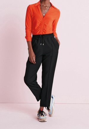 Mujer con una blusa naranja fruncida con botones, pantalones negros de cintura elástica, collares dorados en capas y zapatillas beige con azul sobre un fondo rosa pálido.