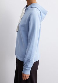 Felpa blu chiaro realizzata in tessuto morbido, con tasca a marsupio e coulisse bianca. I dettagli delle cuciture arricchiscono il design casual.