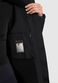 Geacă puffer neagră, cu interior, având un buzunar cu fermoar și o etichetă de îngrijire cu text. Materialul pare neted, cu o finisare mată.