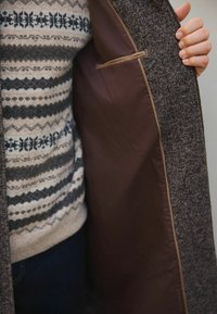 Grå struktureret frakke med en brun foring, der har en skjult lomme. En mønstret trøje er synlig under frakken.