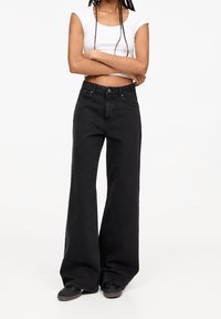 Personne les bras croisés portant un crop top blanc, un jean noir à large jambe et des baskets noires sur un fond blanc uni.