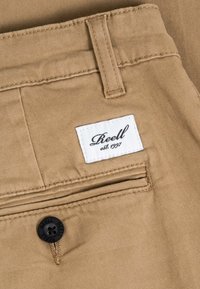 Primer plano de pantalones color beige que muestran un bolsillo trasero con un botón negro y una etiqueta rectangular blanca que dice "Reell est. 1997."