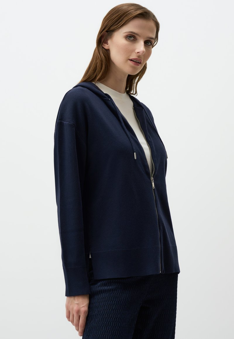 Felpa con zip color navy, in tessuto morbido, vestibilità rilassata, maniche lunghe, due tasche anteriori, dettaglio della zip argento, abbinata a pantaloni scuri con texture.