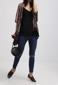 Kimono-style kavaj i mönstrad tyg med röda, svarta och vita inslag. Bärs över en svart topp, matchad med blå slim fit jeans och vinröda slip-ons.