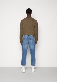 Mężczyzna zwrócony tyłem, ubrany w oliwkowy sweter, jasnoniebieskie krótkie dżinsy, białe skarpetki i białe trampki na jednolitym, jasnoszarym tle.