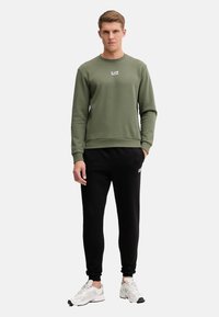 Felpa verde oliva con collo rotondo e piccolo logo bianco, abbinata a pantaloni neri a vita alta e scarpe da corsa bianche.