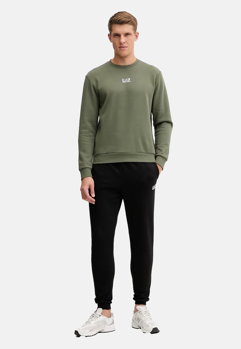 Felpa verde oliva con collo rotondo e piccolo logo bianco, abbinata a pantaloni neri a vita alta e scarpe da corsa bianche.