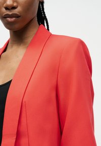 Γυναίκα με πλεξούδες που φοράει ένα λαμπερό κόκκινο σακάκι πάνω από ένα μαύρο τοπ, εμφανίζεται από τους ώμους και πάνω με φόντο ένα απλό φόντο.