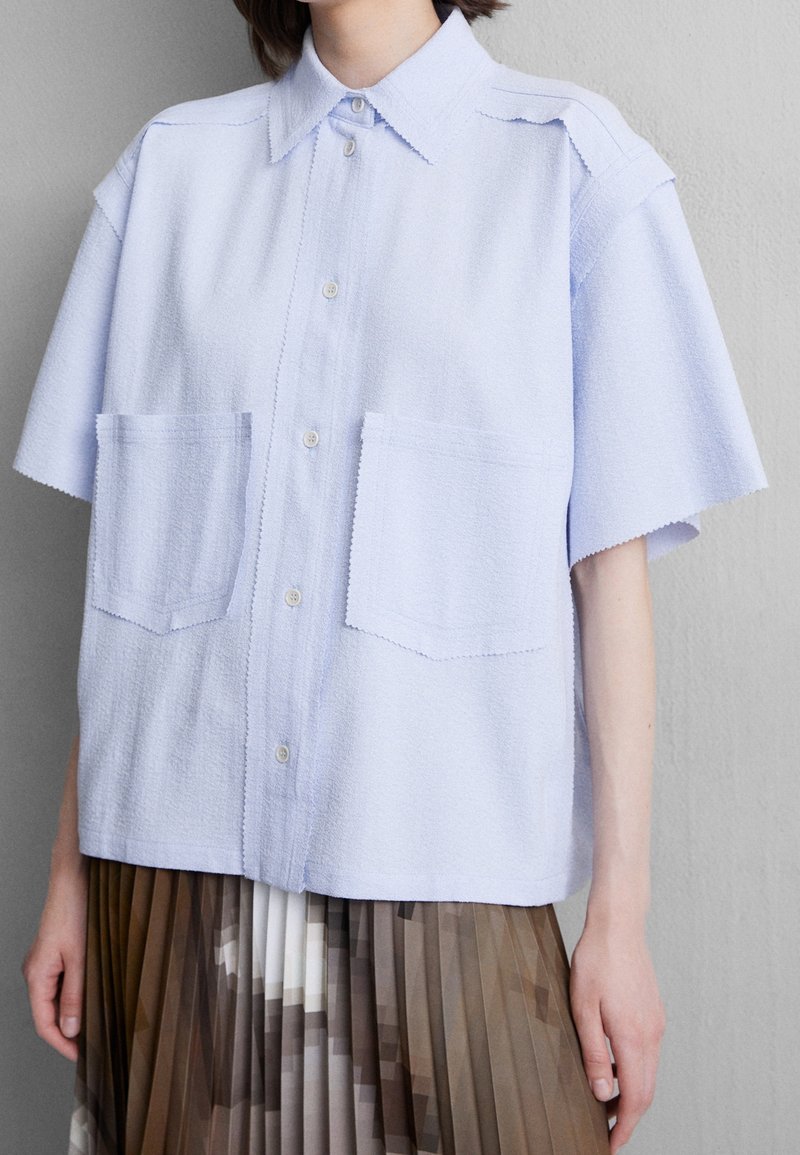 Jasnoniebieska koszula z krótkim rękawem z teksturowanej tkaniny, z zapięciem na guziki i dwiema dużymi kieszeniami naszytymi, zestawiona z brązową spódnicą plisowaną.