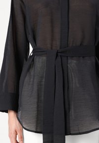 Bluza neagră transparentă cu talie încordată, mâneci lungi și o țesătură texturată. prezintă un croi drept și un luciu subtil.