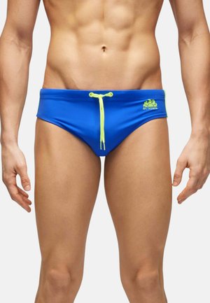 Torso maschile che indossa slip da bagno blu con coulisse gialla e logo Sundek sul lato sinistro, in piedi con le braccia rilassate lungo i fianchi.