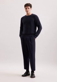 Granatowy sweter ze ściągaczem zestawiony z dopasowanymi granatowymi spodniami. Sweter ma długie rękawy i okrągły dekolt; spodnie mają luźny krój i wyraźne kanty.