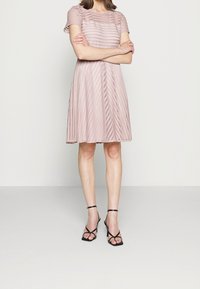 Jasnoróżowa sukienka z krótkim rękawem w poziome paski i diamentowymi wycięciami, rozszerzająca się od talii. Połączona z czarnymi sandałami na wysokim obcasie z paskami.