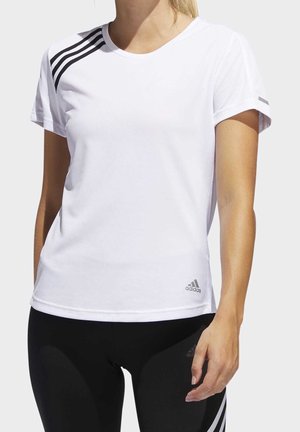 Kvinde iført en hvid Adidas T-shirt med sorte striber på skuldrene og sorte leggings, stående foran en ensfarvet baggrund.
