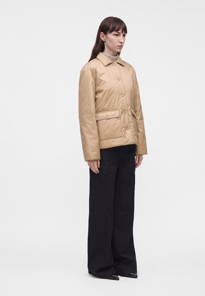 Femme portant une veste matelassée beige par-dessus un pull à col roulé rayé, avec un pantalon large noir et des chaussures noires pointues, debout devant un fond uni.