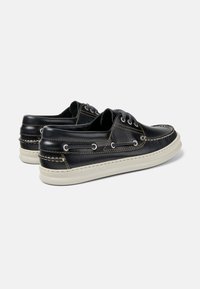 Schwarze Lederbootsschuhe mit weißen Gummisohlen. Merkmale sind weiße Nähte, Schnürdesign und eine niedrig geschnittene Silhouette.