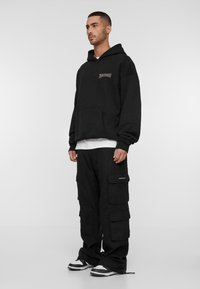 Felpa oversized nera con tasca frontale e logo, abbinata a pantaloni cargo neri con molteplici tasche. Sneakers bianche e nere.